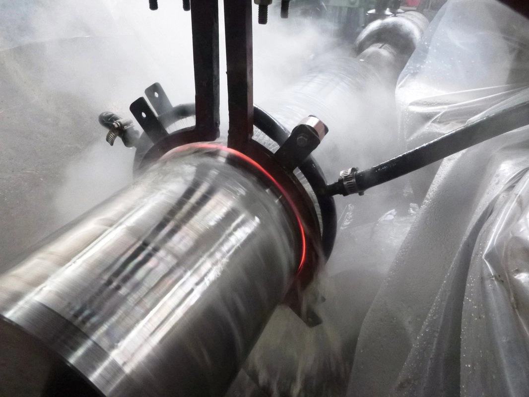 Induction hardening in progress on a rotating steel shaft, a glowing red heat band under the coil and quench mist