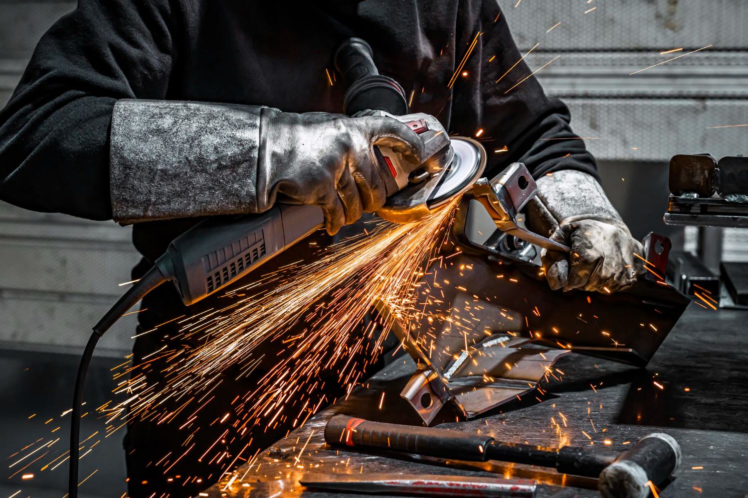 Tradesman grinding a fabricated steel bracket clamped in a vice, a fan of orange sparks across the bench